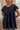 A woman wearing a navy blue short-sleeve tunic top with a ruffled yoke and embroidered detailing, shown in a softly lit indoor setting.