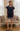 A woman stands in a furnished room wearing a navy blue ruffled short-sleeve top with embroidered detailing, denim shorts, and dark loafers. The room features a vintage-style sofa, patterned cushions, sheer curtains, and a wooden floor.