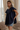 A woman modeling a navy blue short-sleeve top with ruffled layers and embroidered detailing, paired with denim shorts, standing in a softly lit room with curtains and a sofa in the background.