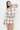 A woman modeling a matching knit cardigan and mini skirt set in a beige and white argyle pattern against a plain light gray studio background.
