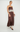 A woman models a brown satin maxi dress with white floral lace trim at the bust and hem, styled with brown heels against a plain light gray studio background.