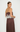 A woman modeling a brown satin maxi dress with a white floral lace overlay at the waist, shown from a rear three-quarter angle against a plain white studio background.
