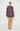 A model stands against a light gray studio background wearing a long-sleeve, oversized knit sweater with horizontal multicolor stripes in purple, blue, pink, orange, and brown, paired with tan knee-high suede boots.