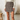 A model wearing a brown and beige plaid wrap mini skirt with a gold-tone ring detail at the waist, paired with a cream long-sleeve top. The setting is a softly lit, modern interior with a textured white wall, a wooden stool, and a white toilet in the background.