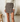 A model wearing a brown and beige plaid wrap mini skirt with a gold-tone ring detail at the waist, paired with a cream long-sleeve top. The setting is a softly lit, modern interior with a textured white wall, a wooden stool, and a white toilet in the background.