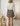 A woman shown from behind wearing a cream long-sleeve top and a brown plaid mini skirt, standing in a softly lit room with a wooden stool and ceramic jug on the left.