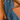 A woman wearing a sleeveless, scoop-neck blue linen dress with side pockets, accessorized with gold jewelry and a woven straw bag, set against a textured terracotta background.