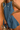 A woman wearing a sleeveless, scoop-neck blue linen dress with side pockets, accessorized with gold jewelry and a woven straw bag, set against a textured terracotta background.