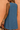 A sleeveless blue shift dress shown from the back on a model, featuring a small keyhole cutout at the neckline with a pearl accent, set against a textured peach background.