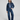 A model stands against a light gray studio background wearing a dark blue cropped denim jacket and matching wide-leg denim jeans over a light blue collared shirt, with brown suede loafers.
