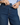 Close-up of a person wearing dark blue denim jeans, pulling the waistband slightly outward with both hands while wearing a light blue and white striped button-up shirt.