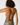 Rear view of a woman wearing a strappy one-piece swimsuit with a beige, black, and gray camouflage pattern, set against a plain light gray background.