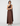 A woman modeling a floor-length, one-shoulder satin dress in a rich dark brown color, standing against a clean, light gray studio background.