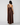 A woman viewed from the back wearing a brown one-shoulder maxi dress with a draped neckline and a long flowing skirt, photographed against a light gray studio background.