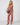 A woman modeling a dusty rose eyelet two-piece set consisting of a sleeveless V-neck top and matching wide-leg pants, styled with a dark brown crescent handbag and black sandals against a light gray studio background.