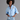 A model with an afro hairstyle wearing a light blue button-down shirt with a pearl and crystal brooch on the chest, paired with a distressed denim mini skirt, against a solid gray studio background.