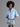 A model with an afro hairstyle wearing a light blue button-down shirt with a pearl and crystal brooch on the chest, paired with a distressed denim mini skirt, against a solid gray studio background.