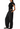 A model wearing a black draped short-sleeve crop top with sheer mesh shoulder panels and matching black wide-leg pants, paired with black flats, against a solid white background.