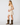 A woman modeling a short cream lace slip dress with spaghetti straps and brown cowboy boots, standing against a light gray studio background.