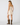 A woman modeling a cream lace slip dress with spaghetti straps and tie-back details, paired with brown cowboy boots, shown from a rear three-quarter view against a light gray studio background.