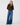 A woman modeling a brown knit zip-up cardigan with a textured collar, paired with wide-leg blue jeans, against a plain light gray studio background.