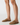 A close-up of a person's legs wearing tan suede mule loafers against a clean white studio background.