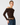 A woman modeling a dark brown off-the-shoulder long-sleeve crop top with blue jeans and a brown belt, photographed against a light gray studio background.