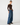 A model stands against a light gray studio background wearing a dark brown off-the-shoulder long-sleeve top, wide-leg blue jeans, a brown belt, and dark brown loafers.