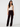 A model stands against a light gray studio background wearing dark burgundy velvet wide-leg pants, a white ribbed tank top, and black shoes.