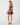 A woman modeling a short sleeveless dress with a floral print in purple, pink, and green tones, shown from the back against a light gray studio background.