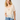A woman wearing a sheer cream ruffled blouse with lace trim and blue jeans, standing against a plain light gray studio background.