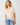 A woman wearing a sheer cream ruffled blouse with lace trim and blue jeans, standing against a plain light gray studio background.