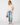 A model stands against a light gray studio background wearing a cream lace-trim blouse with puff sleeves, distressed light-wash blue jeans, beige flats, and holding a small brown handbag.