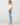 A woman modeling a white ruffled short-sleeve blouse and light-wash distressed jeans with rolled cuffs, paired with beige flat shoes, against a plain light gray studio background.