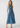 A woman modeling a sleeveless, high-neck, midi-length blue denim dress against a plain light gray studio background.