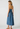 A woman modeling a blue denim midi dress from the back, featuring a tie-shoulder design and an open back, set against a plain light gray studio background.