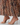 A close-up of a person wearing nude beige strapless high-heel sandals with a pleated skirt featuring a brown and beige spotted pattern.