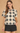 A woman modeling a short-sleeve crochet knit top with a black and beige geometric diamond pattern, lace-up keyhole neckline, and ruffled trim, paired with black shorts against a textured beige background.
