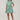 A woman modeling a short tiered mini dress with vertical green, blue, and white stripes, paired with white slide sandals, against a plain light beige studio background.