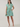 A woman modeling a short tiered mini dress with vertical green, blue, and white stripes, paired with white slide sandals, against a plain light beige studio background.