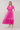 A woman modeling a bright pink midi dress with short puff sleeves, a gathered waist, and embroidered floral details, paired with white heeled sandals against a plain beige studio background.