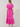 A woman viewed from the back wearing a bright pink midi dress with short puff sleeves, a gathered waist, and embroidered floral motifs, photographed against a plain light beige studio background.