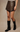 A model wearing a dark brown faux-leather mini skirt and black knee-high leather boots, standing against a neutral beige studio backdrop.