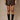 A model wearing a dark brown faux-leather mini skirt and black knee-high boots, standing against a neutral beige studio backdrop.