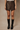 A model wearing a dark brown faux-leather mini skirt and black knee-high boots, standing against a neutral beige studio backdrop.