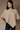 A woman modeling a beige textured knit poncho sweater with a geometric diamond pattern, paired with dark blue jeans, against a neutral brown studio backdrop.