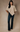 A woman modeling a beige textured knit poncho top paired with dark blue flared jeans and white pointed-toe heels, standing against a solid warm beige studio backdrop.