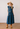 A woman stands in a studio wearing a sleeveless cropped top and matching tiered midi skirt in a dark blue patterned fabric, with a wide-brim straw hat, tan boots, and a long beaded necklace.