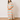 A woman modeling a long-sleeve maxi dress with a colorful pastel checkerboard pattern, standing on a light wood floor against a plain off-white wall.