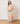 A woman modeling a long-sleeve maxi dress with a colorful pastel checkerboard pattern, standing on a light wood floor against a plain off-white wall.